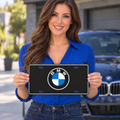 Lifestyle illustration of a model holding the black BMW decorative tag with silver border and blue-and-white roundel beside a blue car.